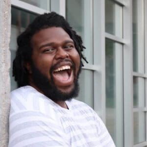 Nik Whitcomb, a Black man with locks wears a gray and white striped shirt while smiling. He is standing in front of a row of windows.